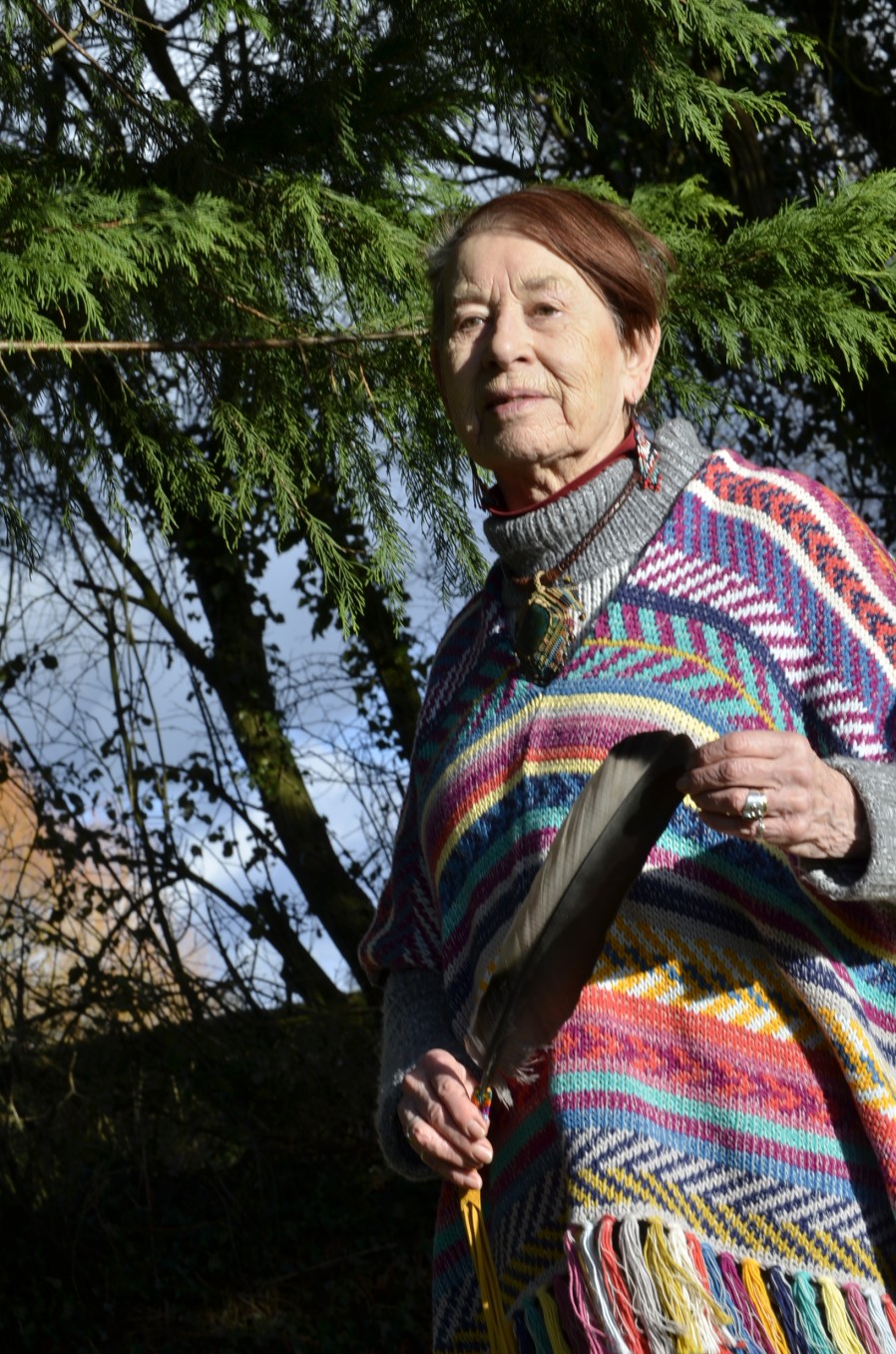 Eliana Harvey, founder of Middle Piccadilly Rural Retreat, wrapped in a rainbow shawl, holding an eagle feather beneath a cedar tree.